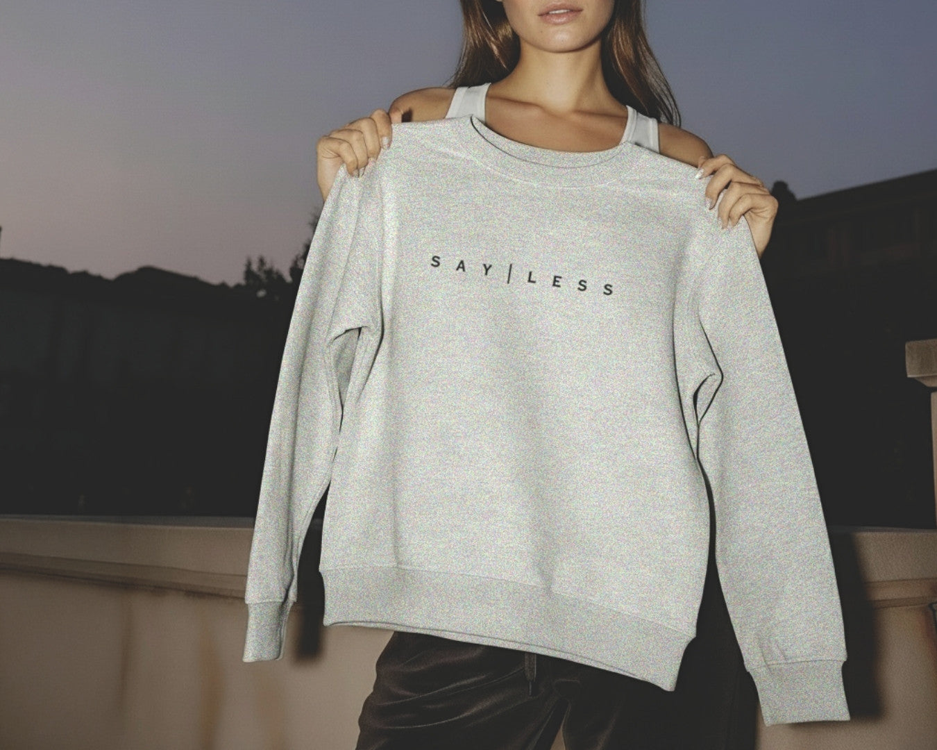 Person holding a gray sweatshirt with 'SAY LESS' text against a blurred outdoor background